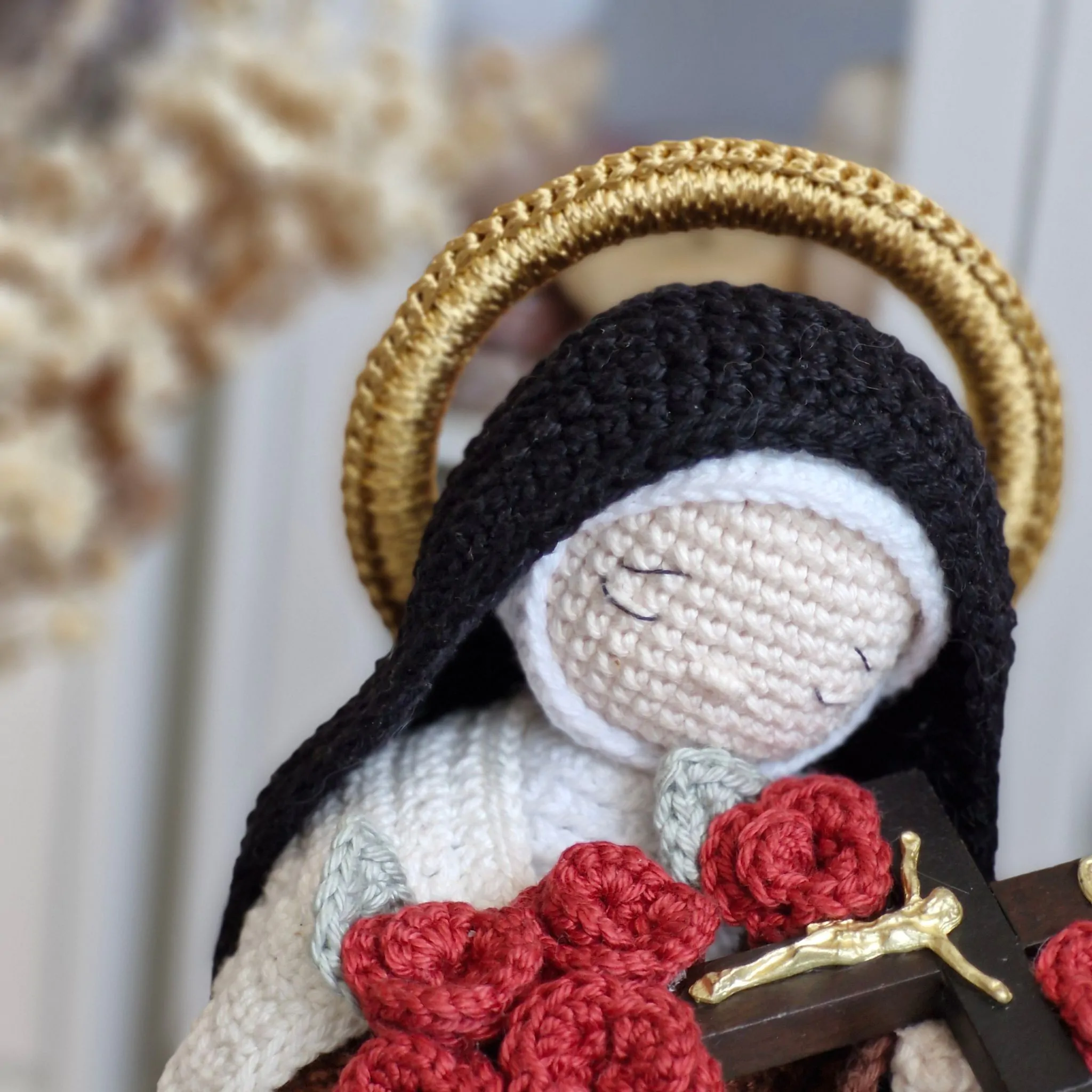 Close da Santa Teresinha do Menino Jesus em amigurumi destacando o rosto bordado, o halo dourado, o crucifixo e as rosas vermelhas.