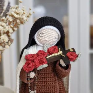 Detalhes da Santa Teresinha do Menino Jesus em amigurumi mostrando o rosto bordado, o crucifixo com rosas vermelhas e o rosário carmelita.