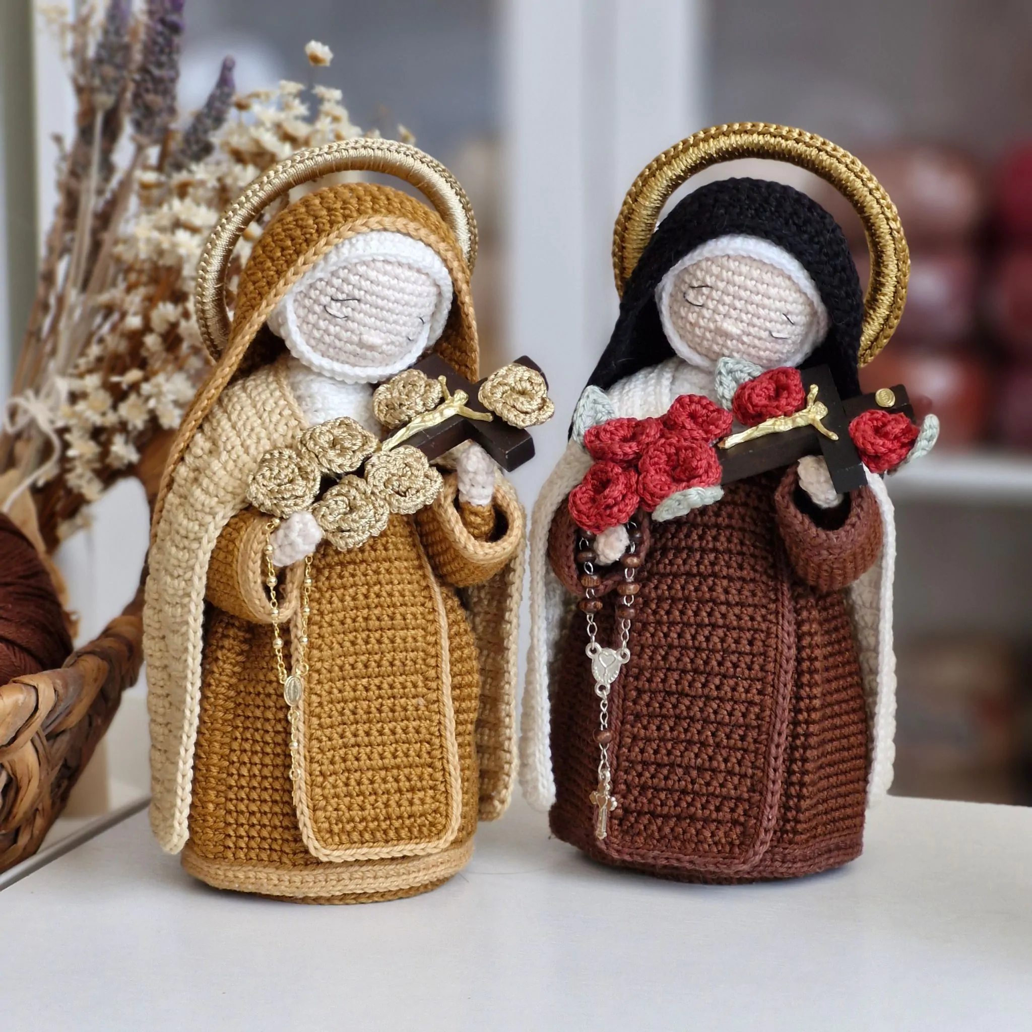 Duas versões da Santa Teresinha do Menino Jesus em amigurumi lado a lado, uma com rosas douradas e outra com rosas vermelhas, ambas segurando crucifixo e rosário.